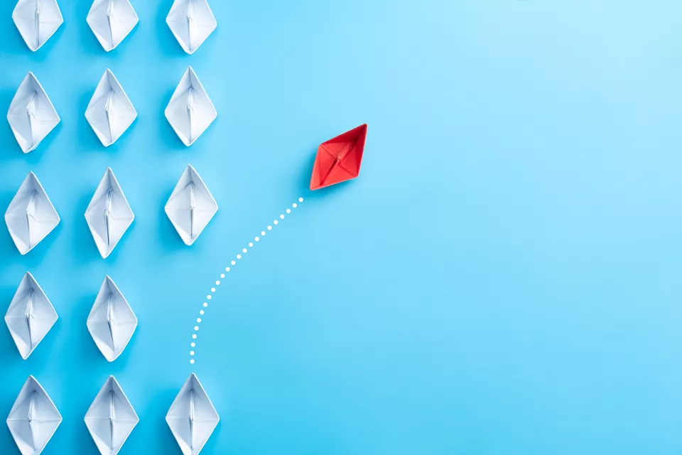 Red paper boat breaking away from white boats, symbolising leadership and innovation.