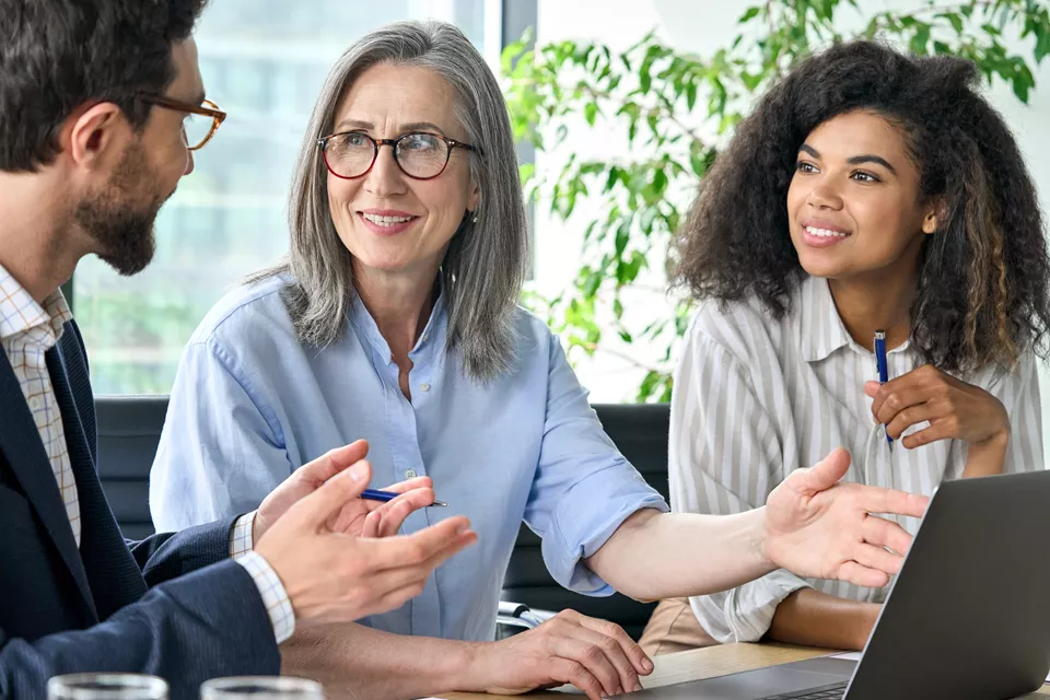 Three people, a man and two women, engaged in a discussion in a professional setting. The woman in the middle, with grey hair and glasses, is smiling and listening attentively. The group appears to be working together on a project, with a laptop open in front of them.