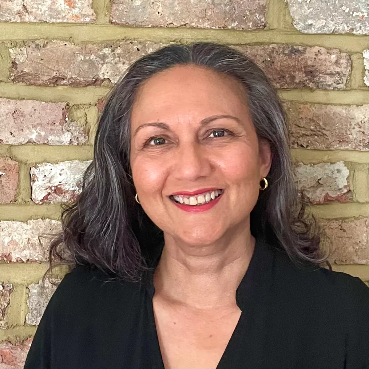 Lubna Gem Arielle - woman with long dark hair and a dark top smiles at the camera. She is stood in front of a brick wall.