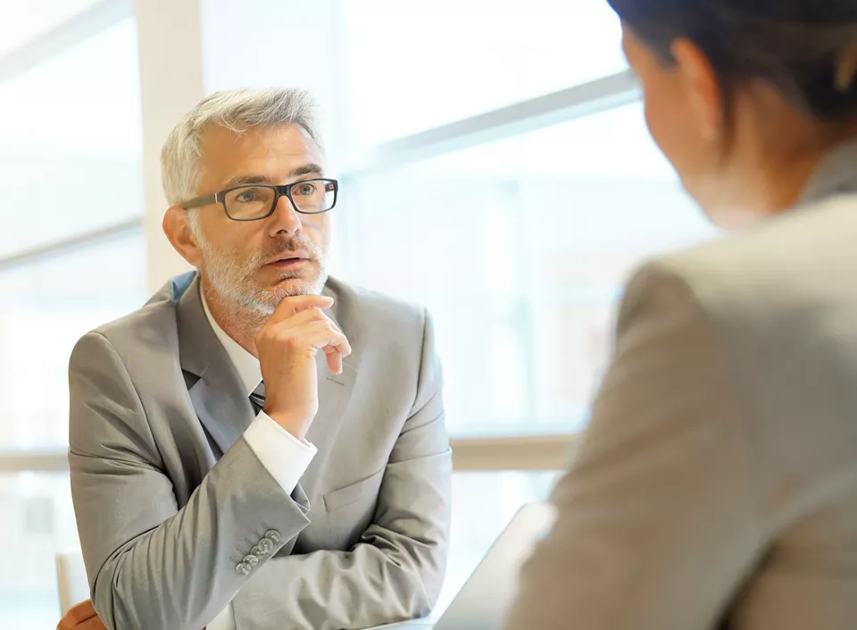 Two professionals in conversation in a bright office setting. A man with grey hair, glasses, and a grey suit is listening thoughtfully, resting his chin on his hand. The back of a second person, also in business attire, is visible in the foreground.