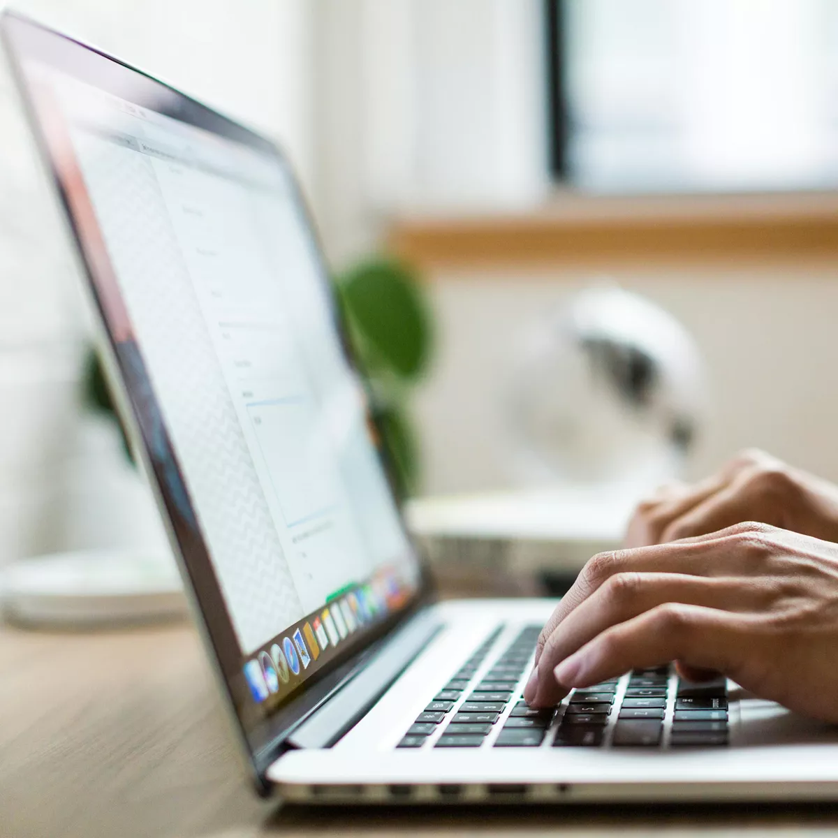 Photo of a man's hands typing into a laptop, viewed from the side. The laptop is on a desk and there is a window in the background, plus an out of focus blue mug and a green plant.