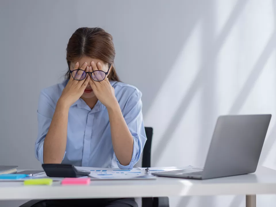 A woman in a blue shirt sits at a desk with her head in her hands, looking stressed or overwhelmed. She is surrounded by a laptop, documents, and office supplies, with sunlight casting shadows on the wall behind her.